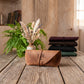 A tan leather roll-up pouch with the letters 'JAL' printed on the front, displayed on a rustic wooden table. In the background, a green fern in a vase and a stack of four folded leather rolls in dark colors (maroon, brown, black, and green) are arranged against a warm, textured wall.