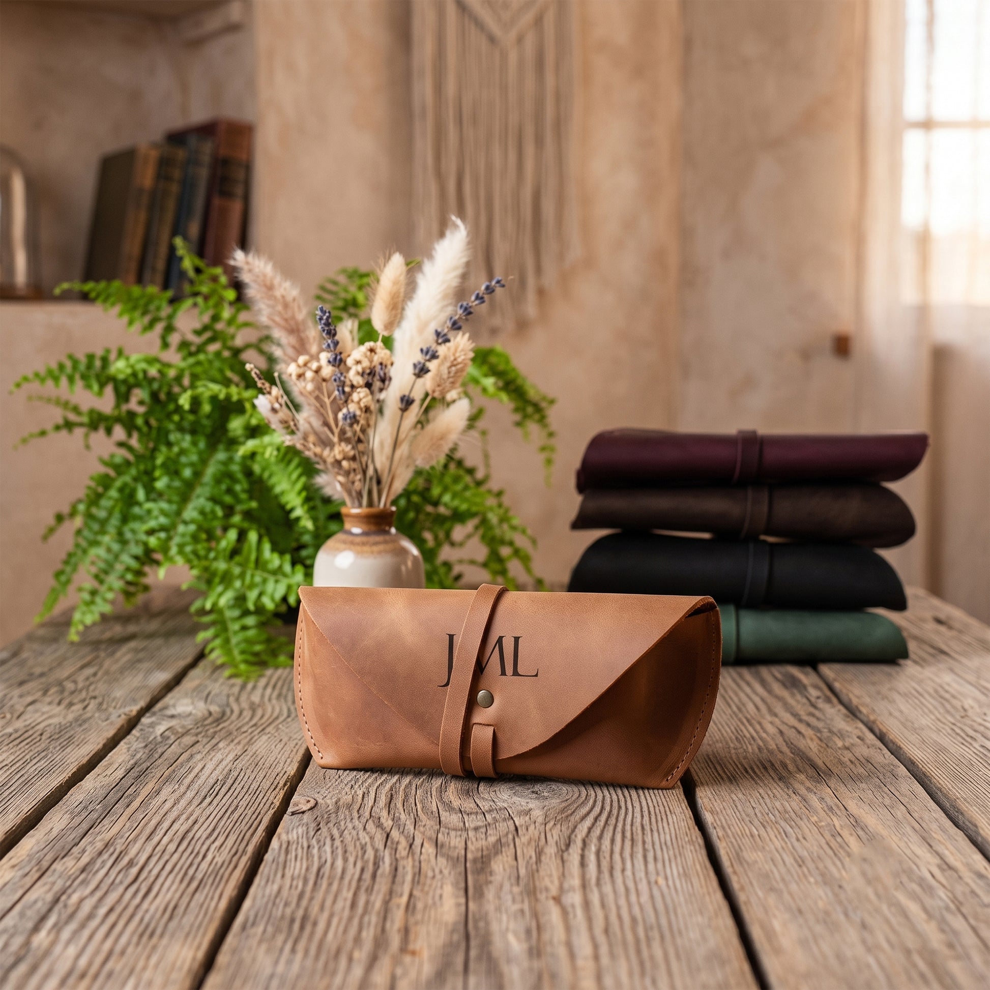 A tan leather roll-up pouch with the letters 'JAL' printed on the front, displayed on a rustic wooden table. In the background, a green fern in a vase and a stack of four folded leather rolls in dark colors (maroon, brown, black, and green) are arranged against a warm, textured wall.