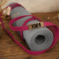 A rolled gray yoga mat secured with a bright pink strap and a brass-toned metal plate engraved with 'YOGA', placed on a wooden surface with dried flowers in the background.
