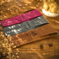 Three personalized leather strap cases in tan, gray, and magenta, laid out diagonally on a rustic wooden surface with dried flowers and warm lighting.