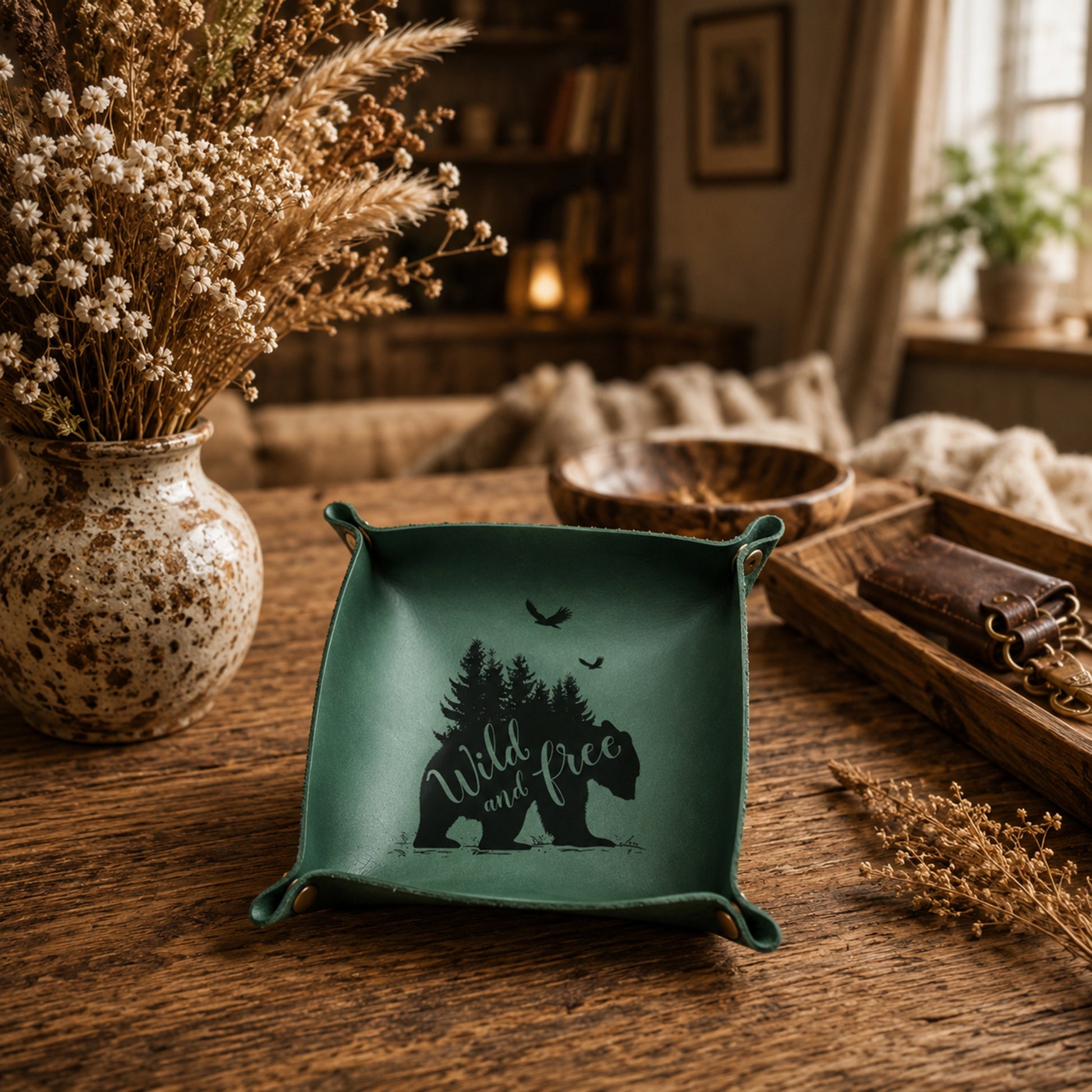 A square teal green ceramic dish with a black graphic of a bear, trees, and the words 'Wild and free' sits on a rustic wooden table in a cozy, warmly lit room. The dish is styled with dried flowers and other home decor in the background.