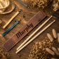 Personalized brown leather drumstick sleeve engraved with the name 'Murphy', displayed on a rustic wooden surface alongside two wooden drumsticks, matching leather wrist straps, and dried floral decor.