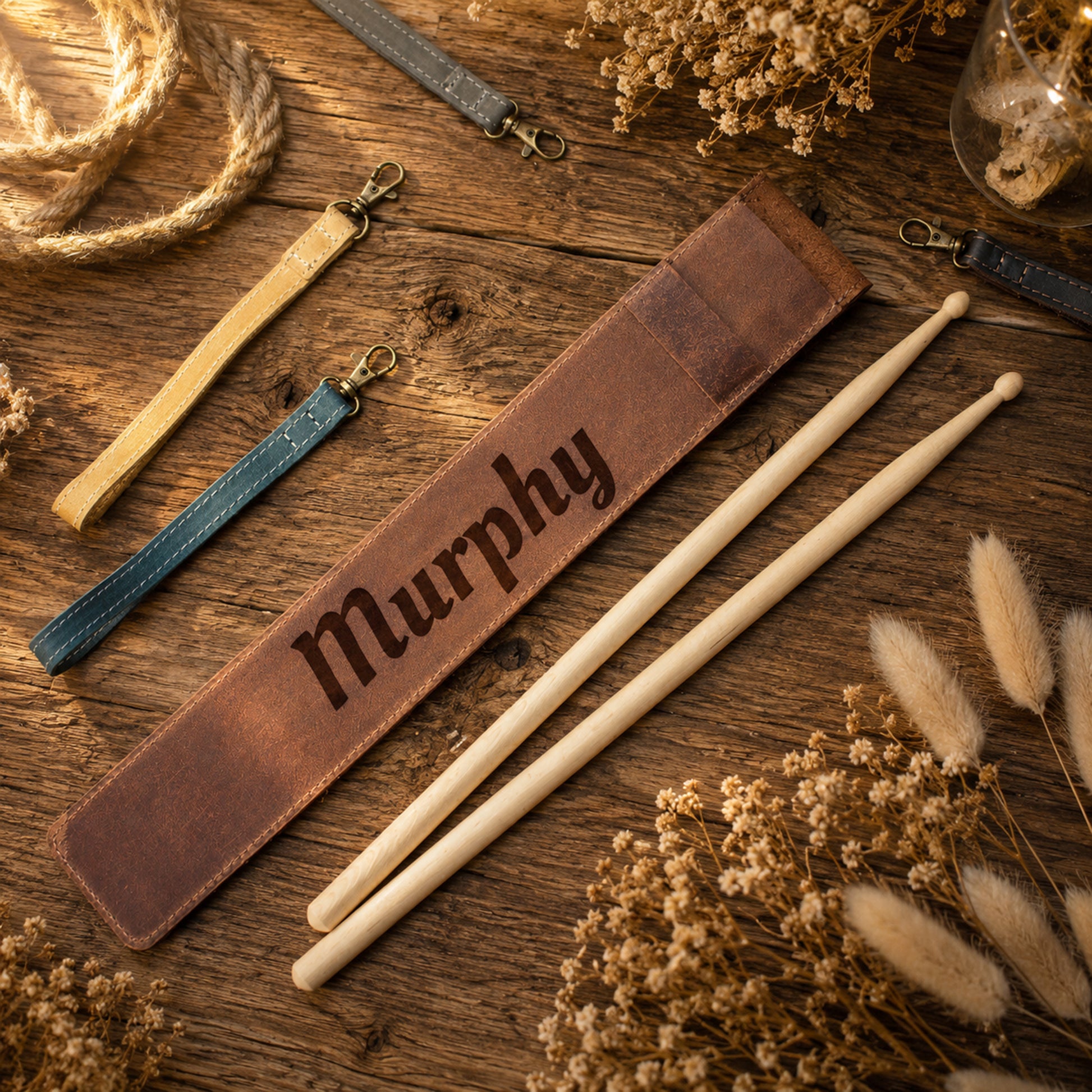 Personalized brown leather drumstick sleeve engraved with the name 'Murphy', displayed on a rustic wooden surface alongside two wooden drumsticks, matching leather wrist straps, and dried floral decor.