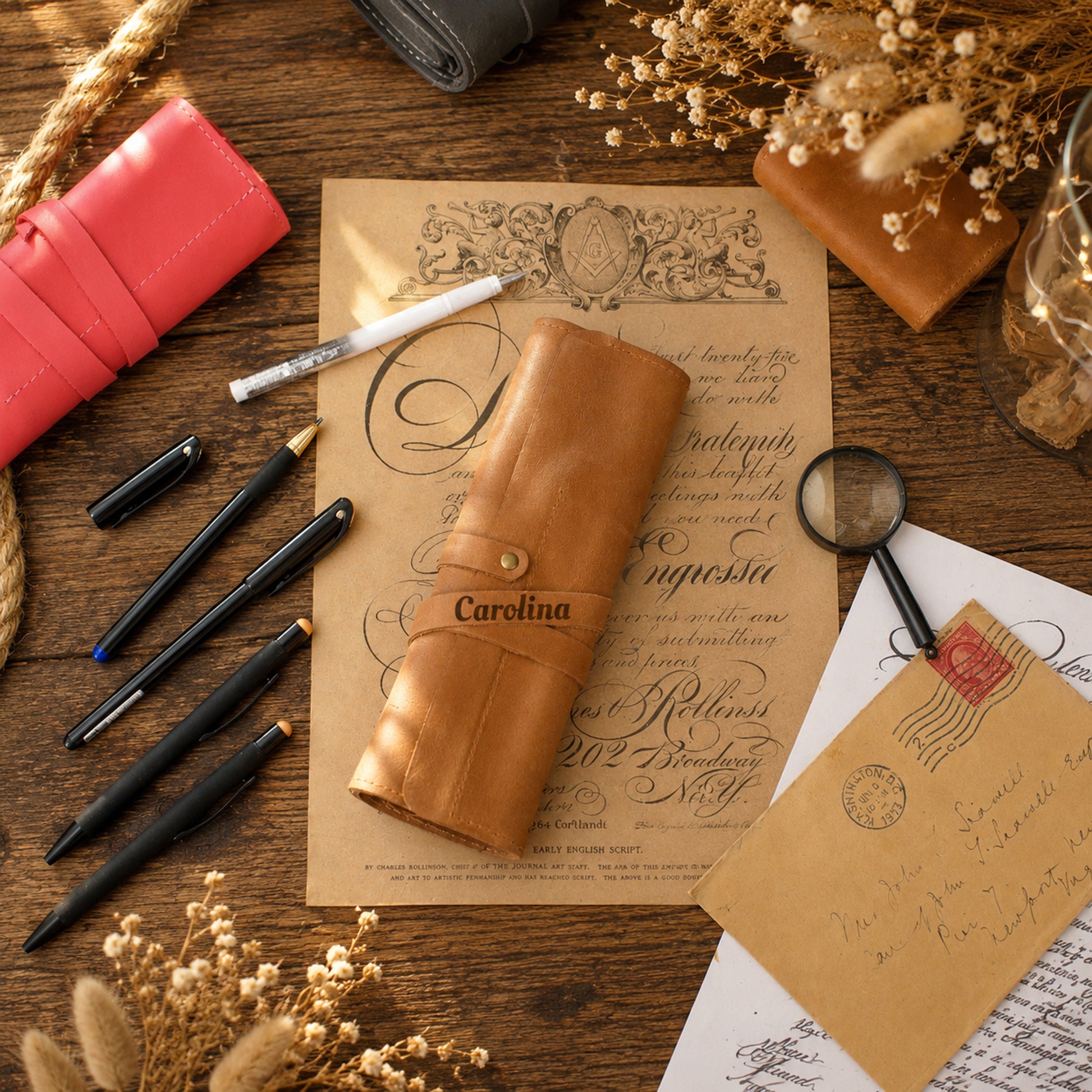 A tan leather pen roll with the name 'Carolina' printed on it, styled in a vintage flat-lay arrangement on a wooden desk with pens, paper, and dried flowers.