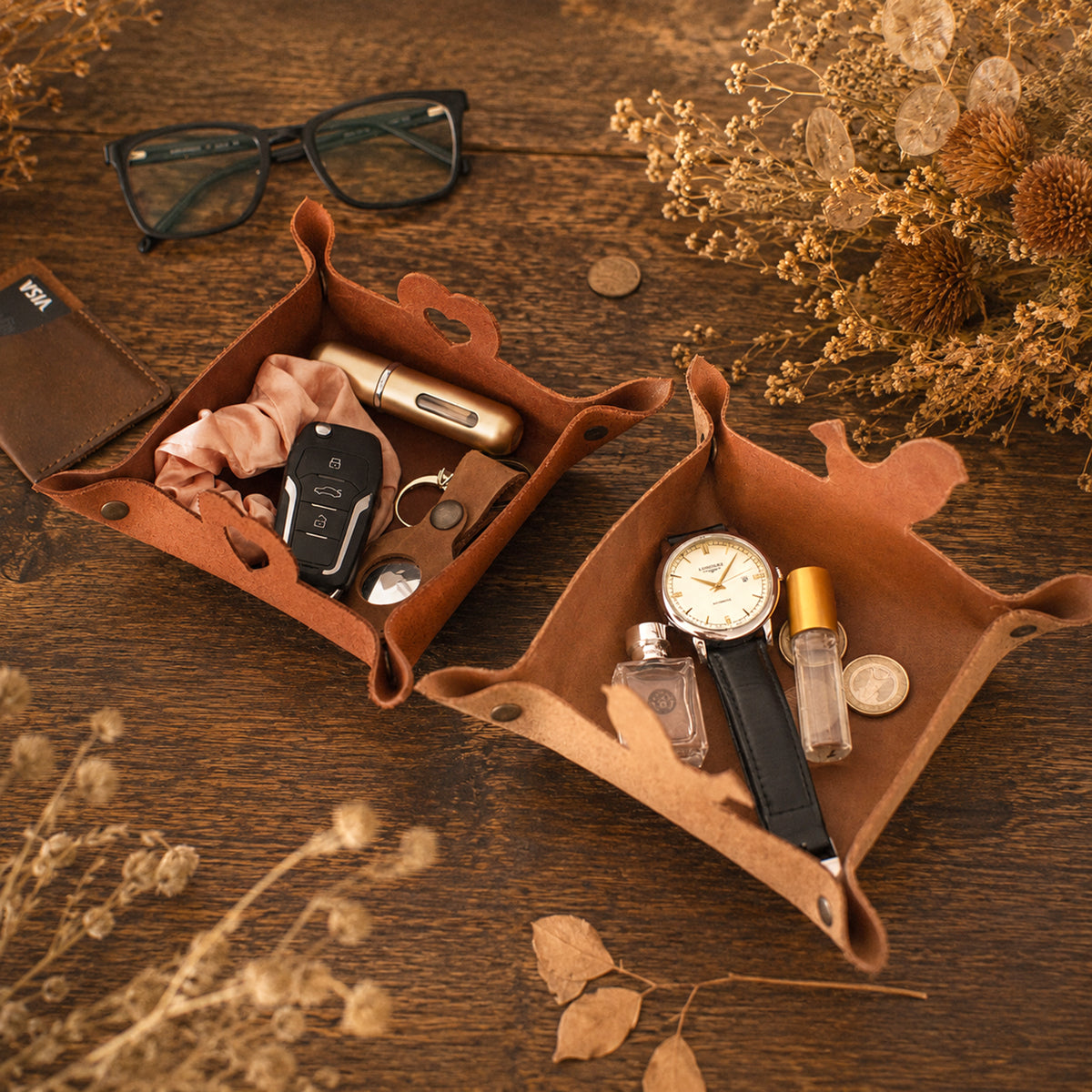 Two brown leather watch and accessory organizer trays arranged on a wooden tabletop with dried flowers, glasses, keys, a watch, perfume bottles, and coins. No person or animal is present, so this is Product/Packaging rather than Lifestyle.