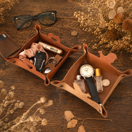 Two brown leather watch and accessory organizer trays arranged on a wooden tabletop with dried flowers, glasses, keys, a watch, perfume bottles, and coins. No person or animal is present, so this is Product/Packaging rather than Lifestyle.