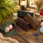 Three personalized leather knitting needle cases in black, brown, and dark green are arranged on a rustic wooden table with knitting props, yarn balls, a small teddy bear, and a lantern in the background.
