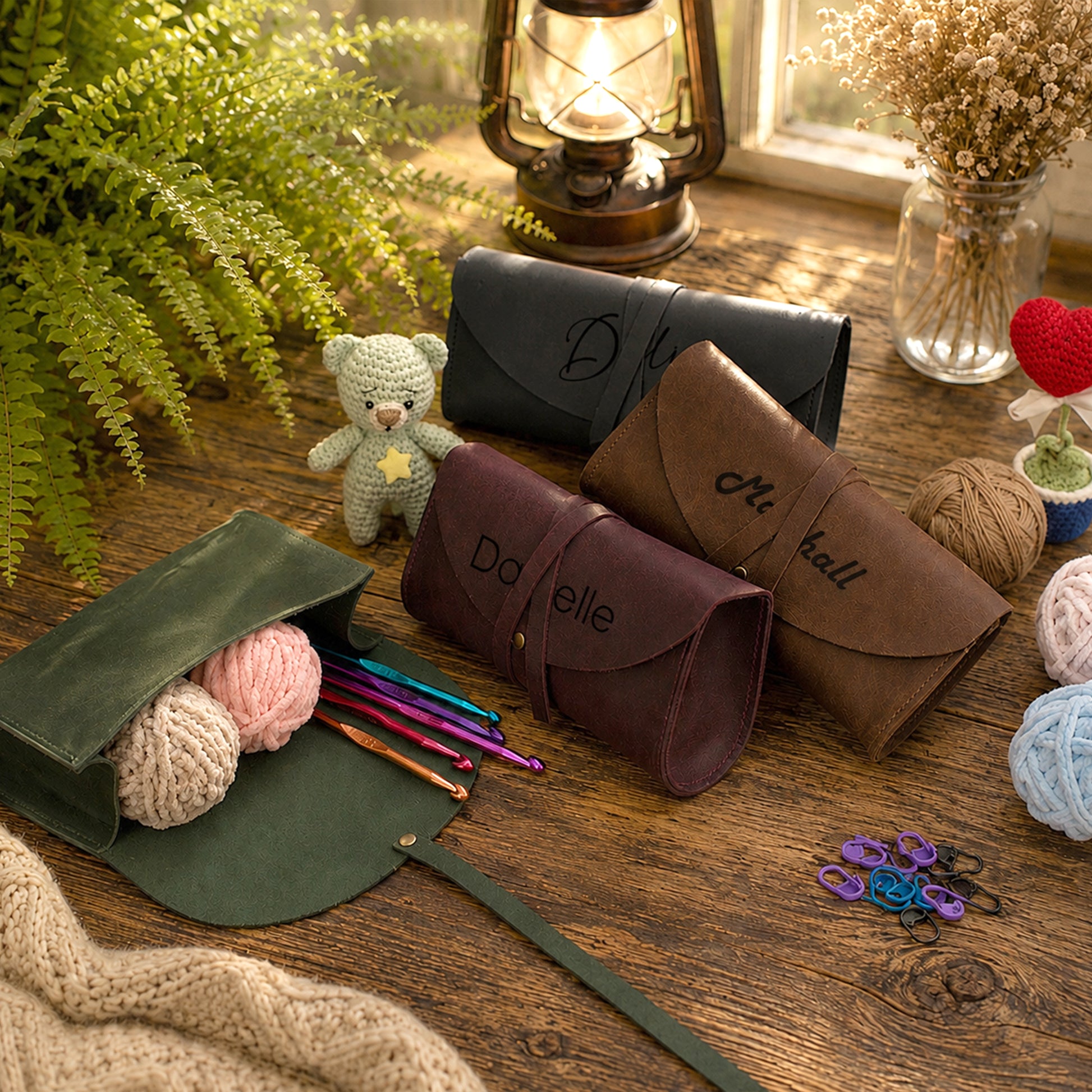 Three personalized leather knitting needle cases in black, brown, and dark green are arranged on a rustic wooden table with knitting props, yarn balls, a small teddy bear, and a lantern in the background.