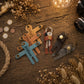 A styled flat-lay of personalized wooden and leather keychains on a rustic wooden table, surrounded by dried flowers, rope, coins, and a camera lens. The keychains include a blue cross with the name 'William', a tan cross with 'DANI', a brown leather key holder, and a dark gray leather key ring.