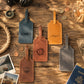 Four personalized leather luggage tags in blue, tan, brown, and gray, styled on a rustic wooden table with travel props like a compass, rope, coins, and a passport.