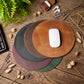 Four round leather mouse pads in brown, gray, mauve, and green are arranged in a stack on a rustic wooden desk, with a white computer mouse on the top brown pad. Decorative props including a keyboard corner, a plant, dried flowers, and earbuds are visible around the setup.
