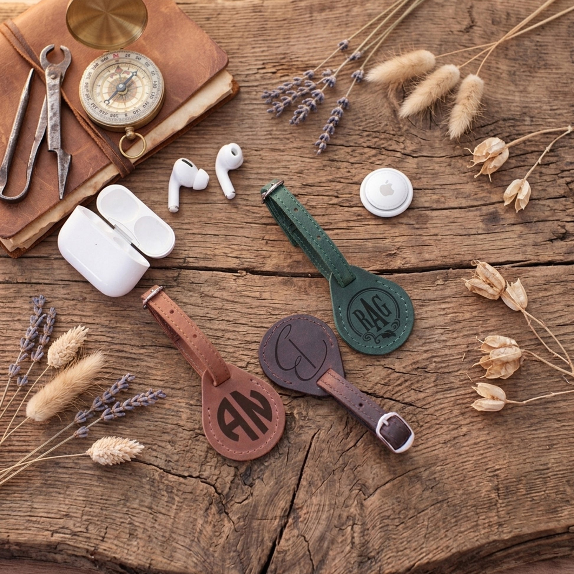 Three personalized leather keychains in brown, dark green, and dark gray are arranged on a rustic wooden table with vintage props including a compass, wrench, book, and dried flowers. White wireless earbuds and a charging case are also present in the flat-lay composition.