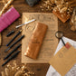 A tan leather pen roll with a snap strap and the name 'Carolina' stitched on the front, styled on a wooden desk with vintage papers, pens, a magnifying glass, and dried flowers.