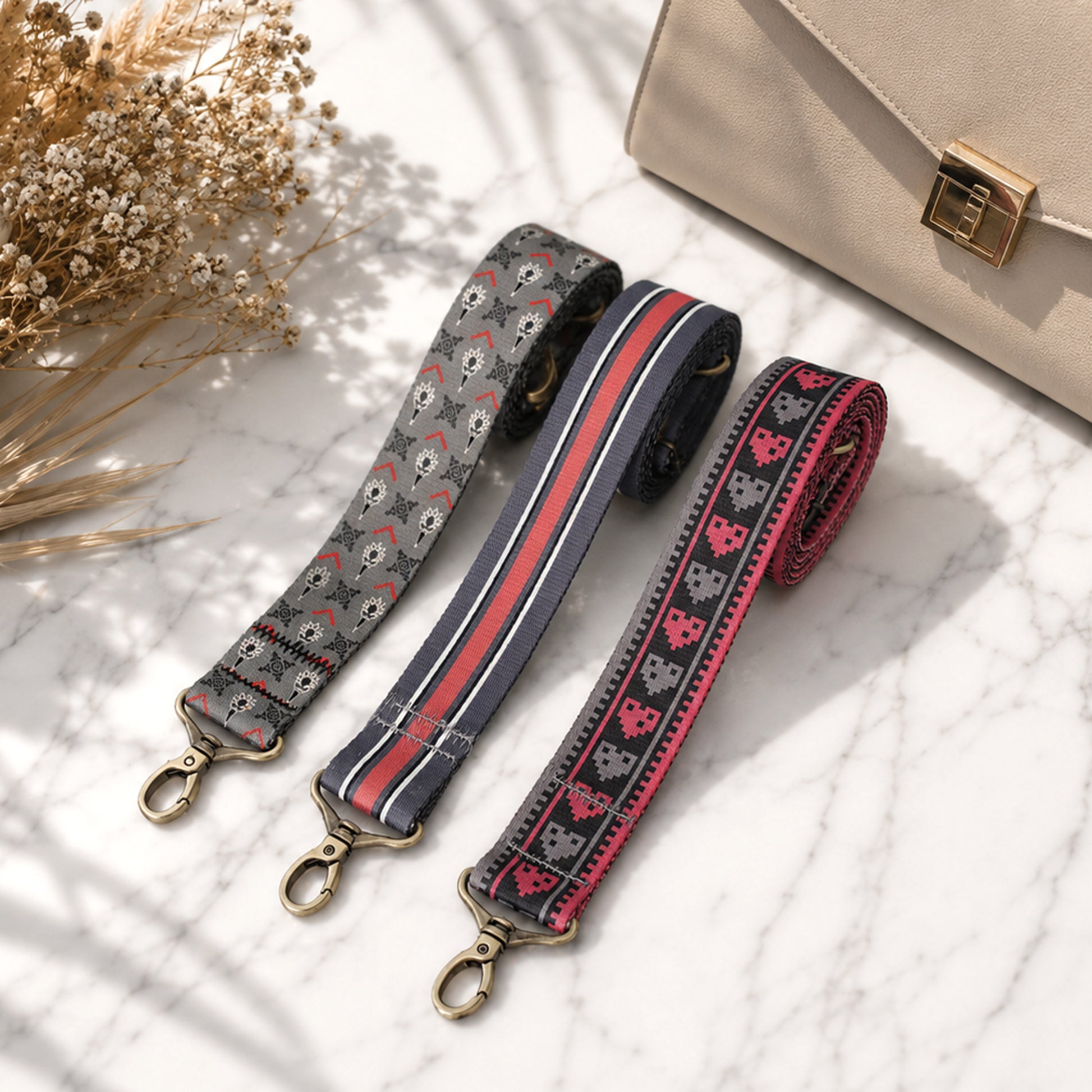 Three patterned bag straps on a light surface with a beige handbag and dried flowers.