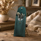 A teal makeup brush pouch with a metal stand, holding several makeup brushes, styled on a rustic wooden table with dried flowers, books, and a beige throw blanket in the background.
