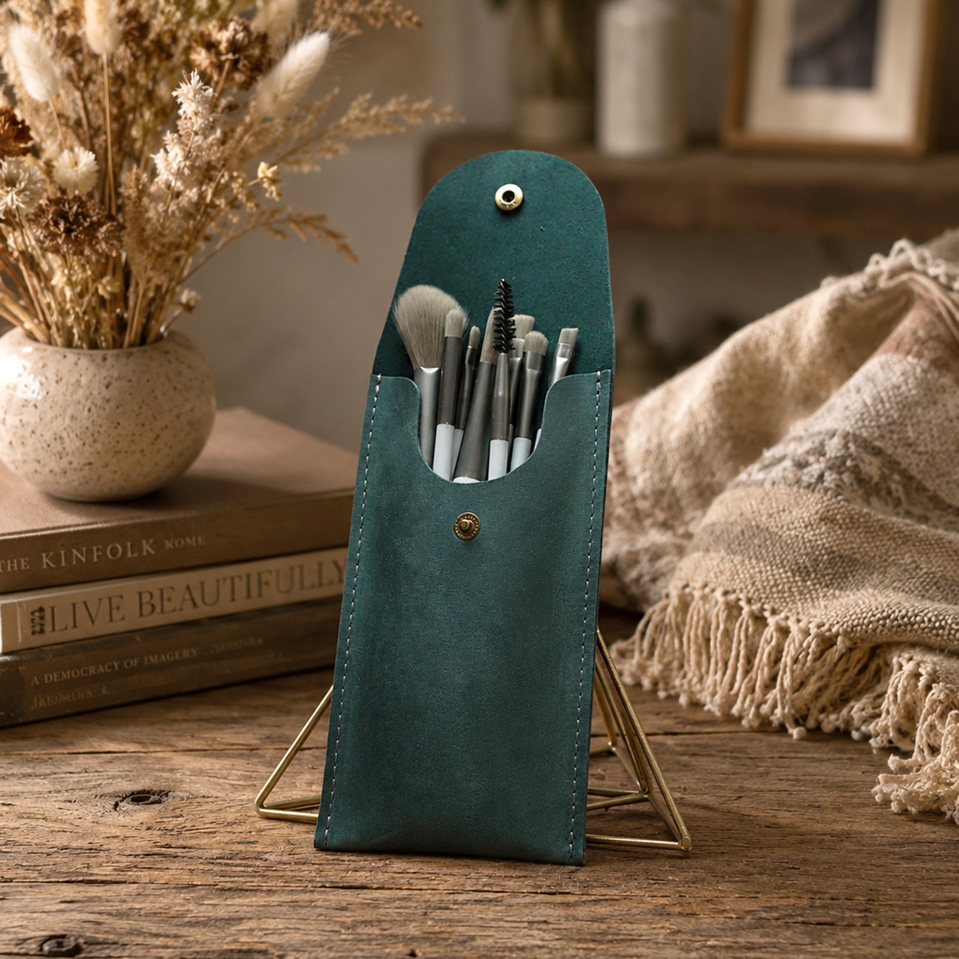 A teal makeup brush pouch with a metal stand, holding several makeup brushes, styled on a rustic wooden table with dried flowers, books, and a beige throw blanket in the background.