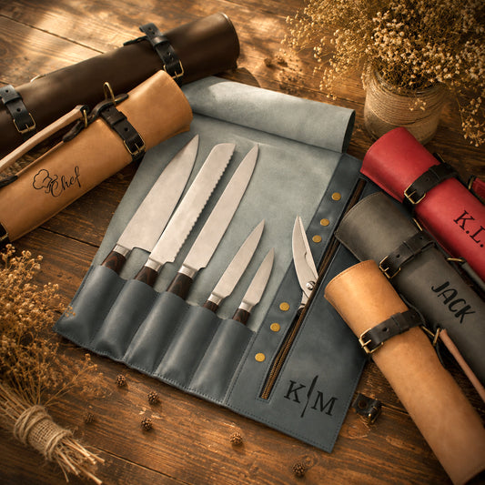 A set of kitchen knives displayed in an open gray fabric roll-up organizer on a wooden table, surrounded by matching rolled knife sleeves in brown, red, and gray. The sleeves are personalized with names such as Chef, K.