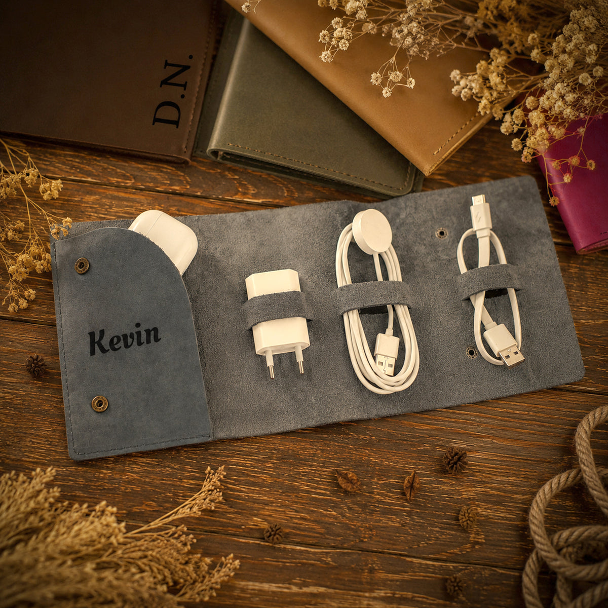 A personalized gray suede roll-up organizer pouch with the name 'Kevin' embroidered on the front, shown open on a rustic wooden surface with dried flowers and accessories inside.