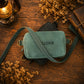 A personalized crossbody camera bag in a muted sage green suede-like material, featuring the name 'WILLIAM' embroidered on the front. The bag is styled in a flat-lay arrangement on a rustic wooden table, surrounded by dried flowers, a vintage lantern, and old books.