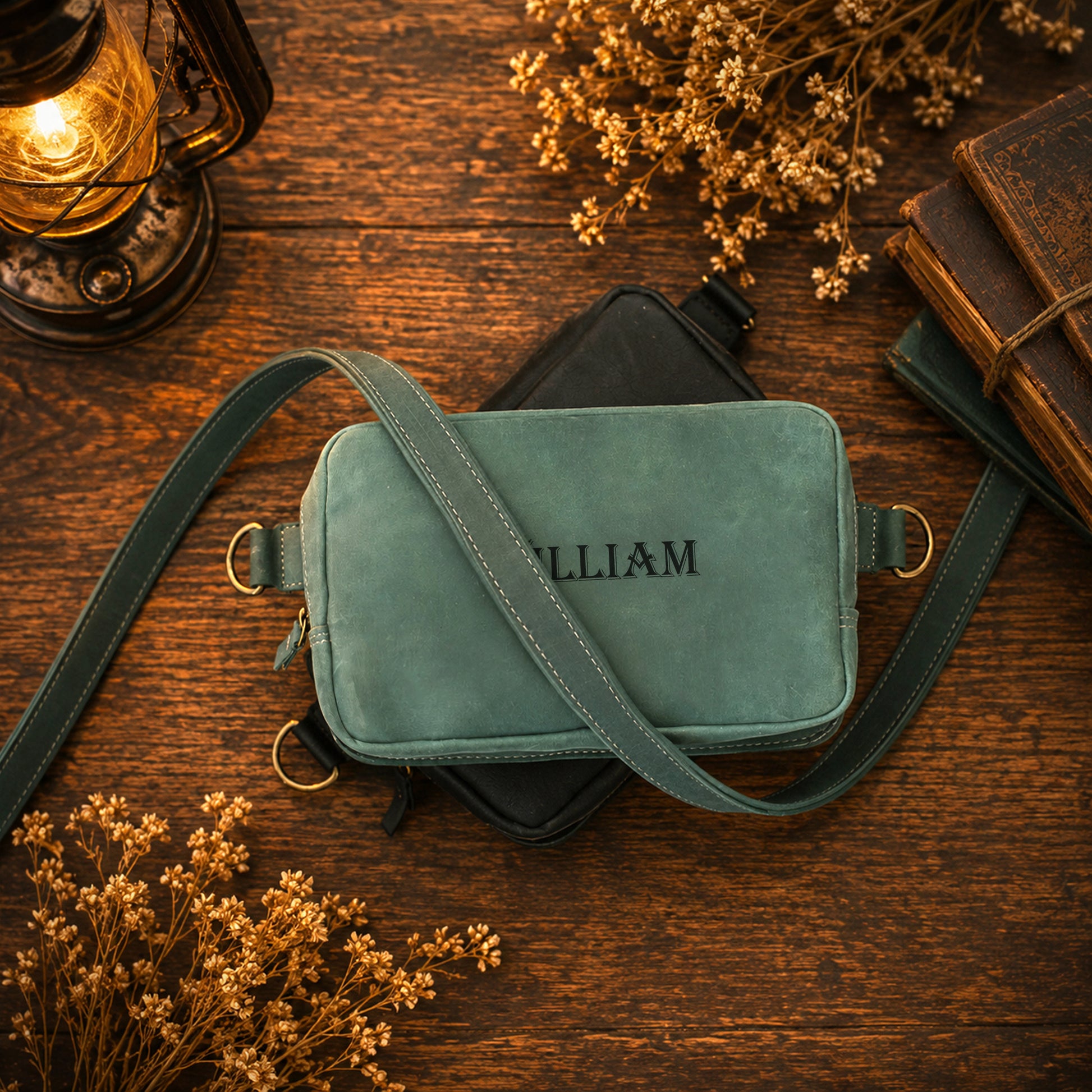 A personalized crossbody camera bag in a muted sage green suede-like material, featuring the name 'WILLIAM' embroidered on the front. The bag is styled in a flat-lay arrangement on a rustic wooden table, surrounded by dried flowers, a vintage lantern, and old books.