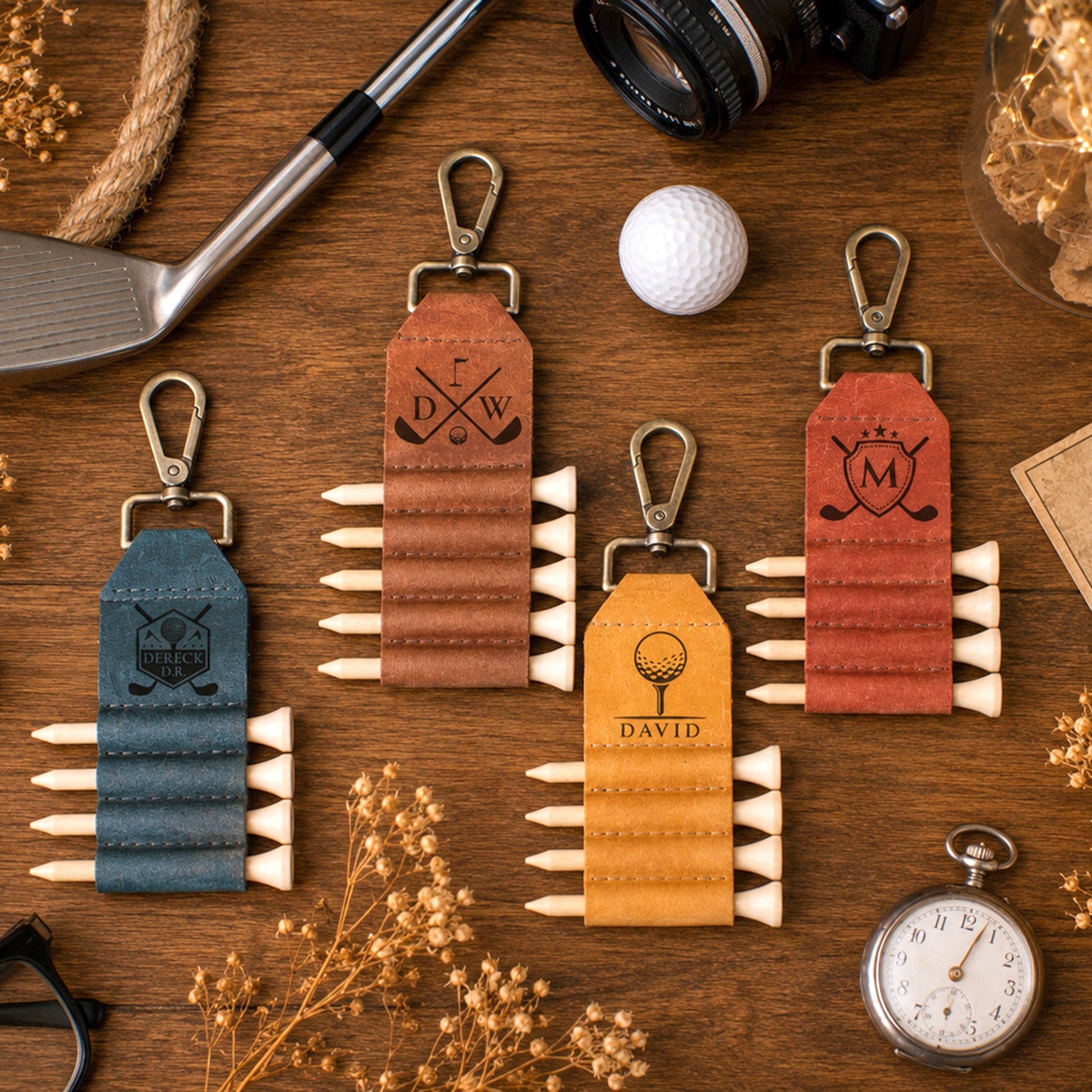 Four personalized golf keychains arranged on a wooden tabletop with golf-themed props. The keychains are made of textured leather in blue, brown, yellow, and terracotta colors, each with metal clasp, a monogram or crest design, and multiple white golf tees attached along the bottom.