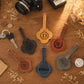 Six personalized leather luggage tags in different colors (tan, brown, black, blue, red-brown, and gray) are arranged on a wooden tabletop with styled props like a camera, keys, and dried flowers. Each tag features a unique monogram or name engraved on a scalloped backplate.