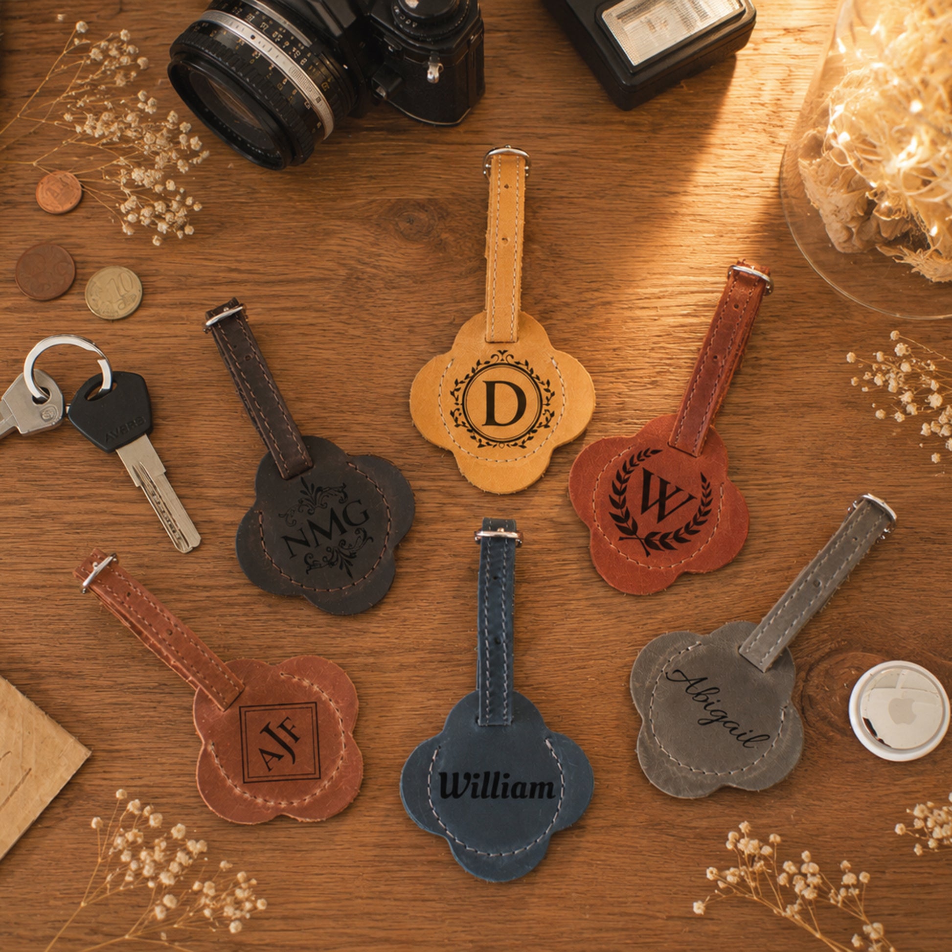 Six personalized leather luggage tags in different colors (tan, brown, black, blue, red-brown, and gray) are arranged on a wooden tabletop with styled props like a camera, keys, and dried flowers. Each tag features a unique monogram or name engraved on a scalloped backplate.