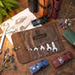 A brown leather tool roll with the brand name 'Marshall' engraved on the front, holding several silver wrenches, arranged on a wooden tabletop with automotive-themed props including toy cars, wrenches, a sketch, and dried flowers.