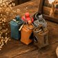 Four small dog poop bags with metal rings and keychains, shown in teal, orange, gray, and brown on a wooden table with dried flowers and a framed dog photo in the background.