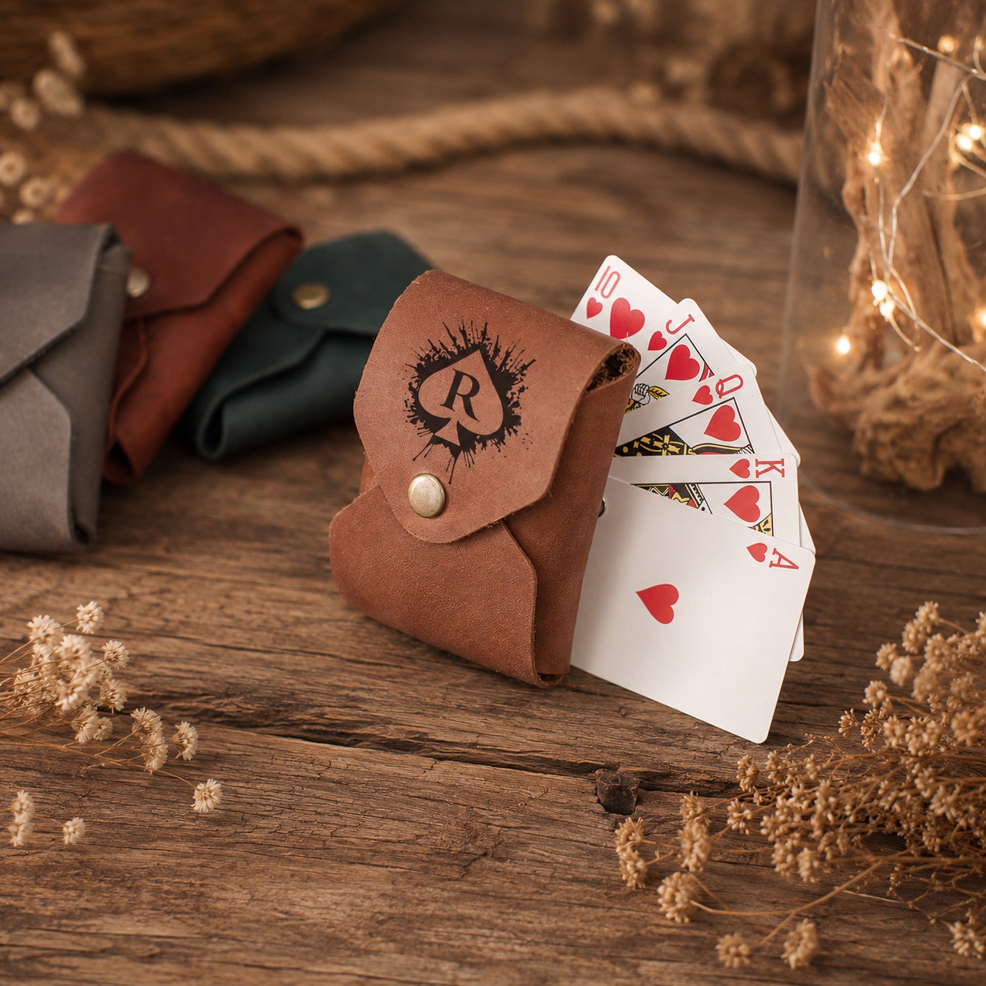 A brown leather card pouch with a snap closure and a black spade logo, shown with playing cards fanned out on a rustic wooden surface. Other pouches in gray and dark green are blurred in the background, along with dried flowers and warm string lights.
