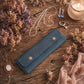 A dark teal blue velvet jewelry wallet with two gold snap buttons, styled on a rustic wooden table surrounded by dried flowers, necklaces, rings, and a lit candle.