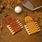 Two leather golf keychains displayed on a wooden surface with a golf ball, club head, and dried flowers. One keychain is reddish-brown leather with a black engraved monogram, and the other is tan leather with an engraved golf club logo.