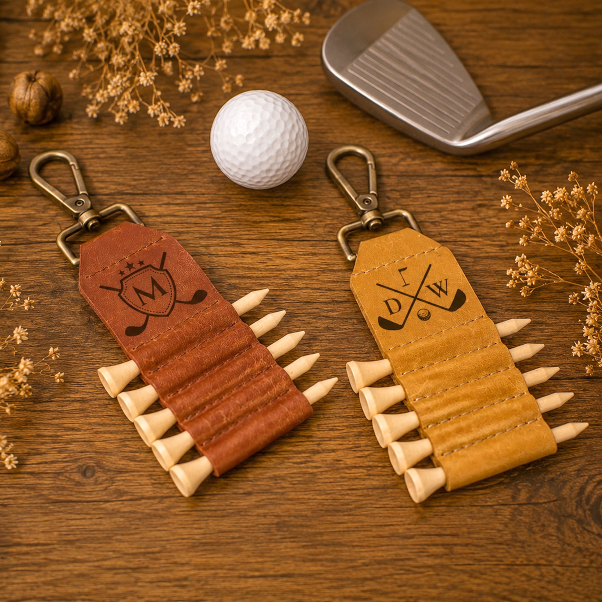 Two leather golf keychains displayed on a wooden surface with a golf ball, club head, and dried flowers. One keychain is reddish-brown leather with a black engraved monogram, and the other is tan leather with an engraved golf club logo.