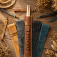 Five personalized drumstick pouches in tan, gray, brown, black, and teal, arranged on a rustic wooden surface with dried flowers and rope. The center brown pouch contains a pair of wooden drumsticks and is personalized with the name 'DANIEL' and a monogram 'D'.