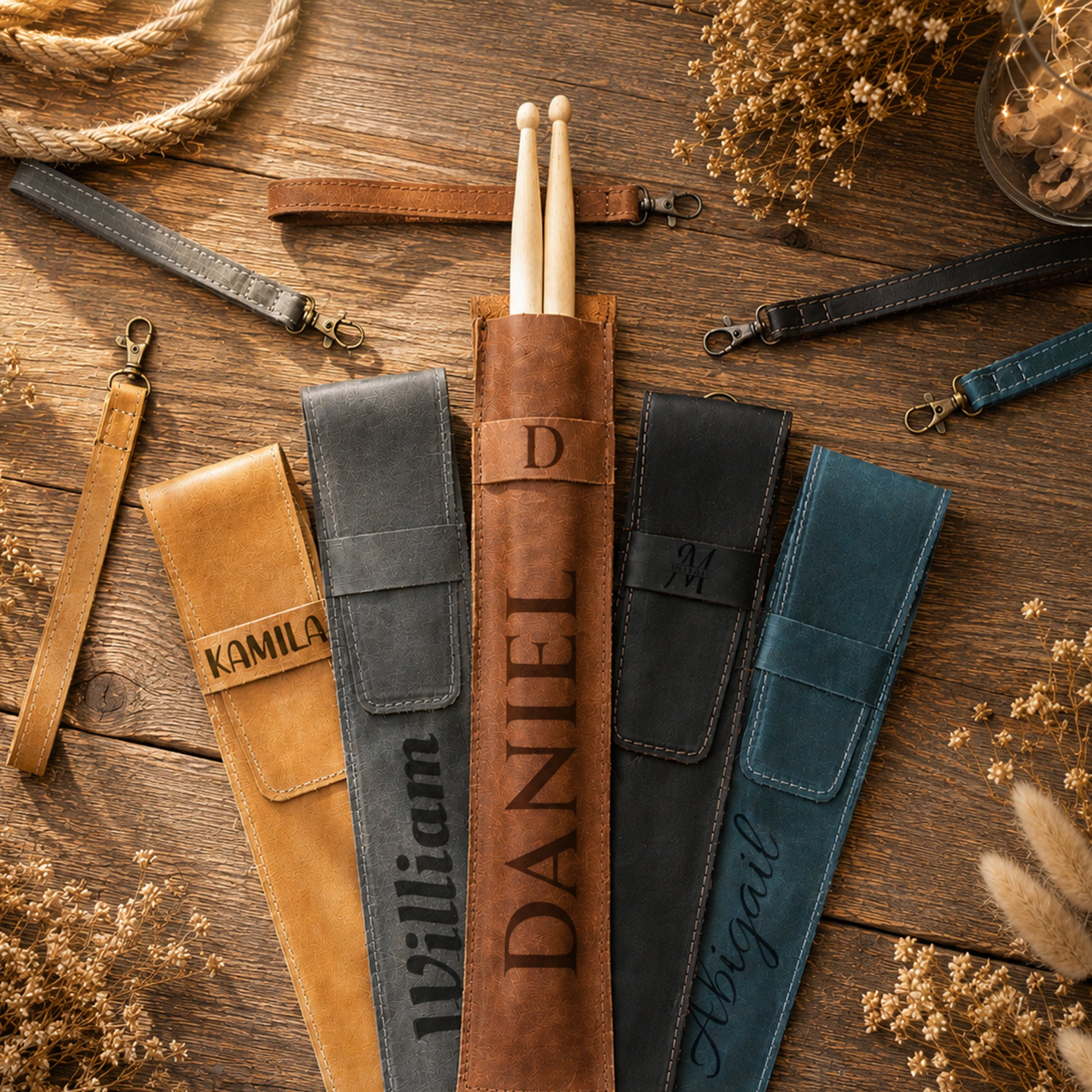 Five personalized drumstick pouches in tan, gray, brown, black, and teal, arranged on a rustic wooden surface with dried flowers and rope. The center brown pouch contains a pair of wooden drumsticks and is personalized with the name 'DANIEL' and a monogram 'D'.