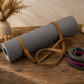 A gray textured yoga mat rolled up with tan straps, displayed on a rustic wooden table next to a coiled black and red strap, with dried flowers in a vase in the background.