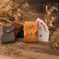 A tan leather card holder envelope with a snap button and an engraved monogram 'A' stands on a rustic wooden surface, fanned with playing cards. A dark gray leather card holder and a brown leather card holder are placed behind it, with dried flowers and warm string lights in the background.