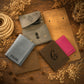 A styled flat-lay of several leather wallets and card holders arranged on a rustic wooden surface with dried flowers, rope, and pinecones. The set includes a light blue wallet engraved with 'JACOB', a brown wallet with a large black monogram, a bright pink folded wallet, a tan card case, and two taupe wallets with snap closures.