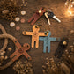 Four leather-shaped keychains arranged on a rustic wooden surface with dried flowers, rope, coins, and a string of warm fairy lights. The keychains are in brown, tan, light blue, and black, each with a metal key ring and a small round snap button.