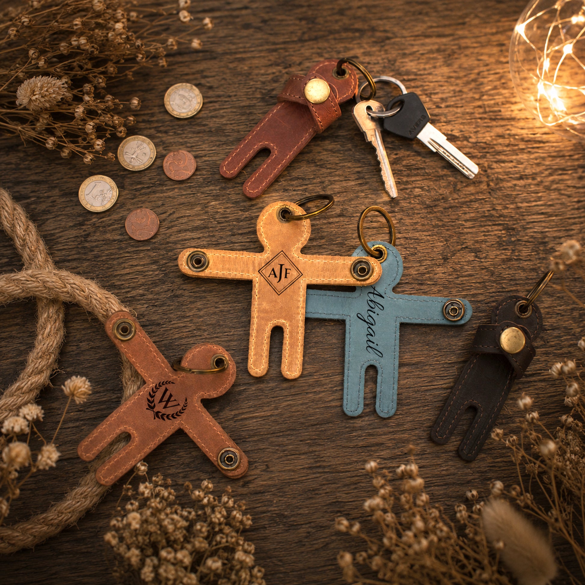 Four leather-shaped keychains arranged on a rustic wooden surface with dried flowers, rope, coins, and a string of warm fairy lights. The keychains are in brown, tan, light blue, and black, each with a metal key ring and a small round snap button.