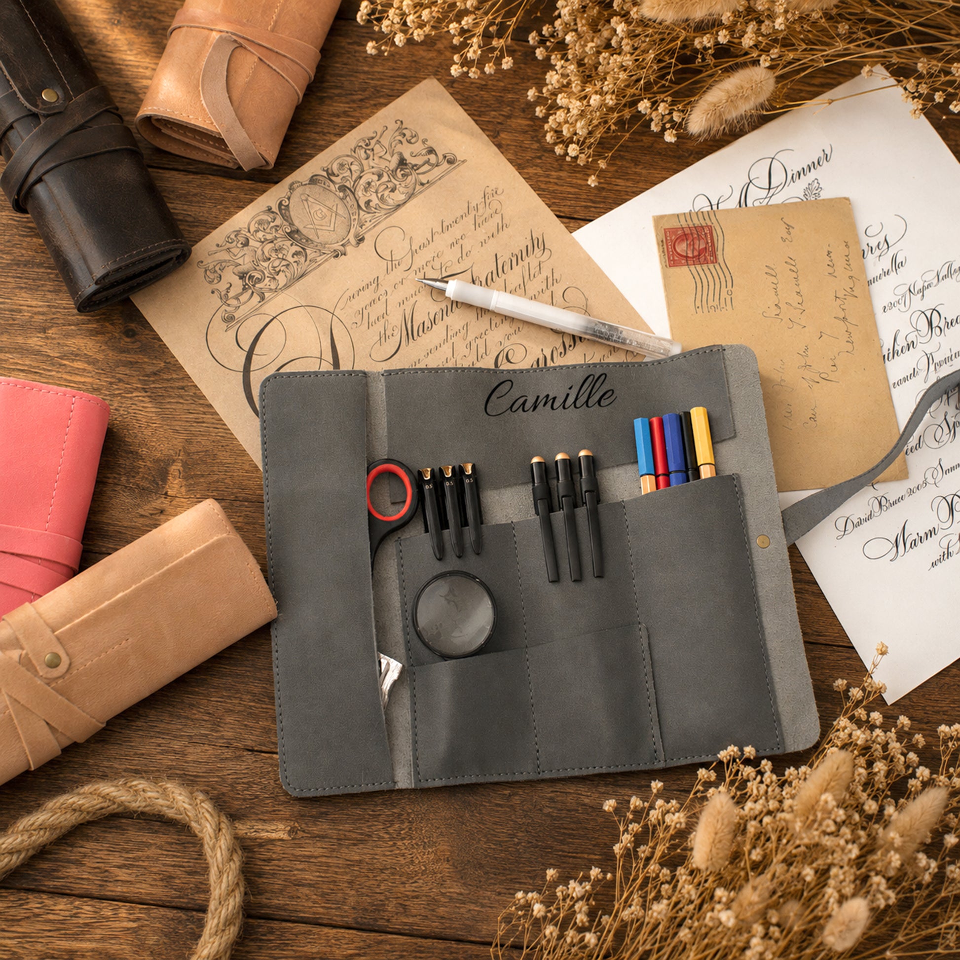 A gray suede wallet organizer laid open on a wooden surface, holding pens, scissors, a magnifying glass, and small accessories. The wallet is styled with stationery, dried flowers, rope, and leather wallets around it.