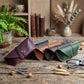 Four leather eyeglasses cases in burgundy, black, tan, and forest green are arranged on a rustic wooden table with decorative props including a potted fern, books, dried flowers, and a pair of eyeglasses.