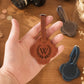 A hand holds a brown leather luggage tag with an engraved monogram 'W' inside a laurel wreath. The background is a wooden tabletop with other leather accessories, including a blue one.