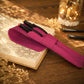 A magenta pink faux-leather pen pouch resting on an open sketchbook on a wooden desk, with two black pens tucked inside. The scene is styled with dried flowers and a warm, glowing glass jar in the background.