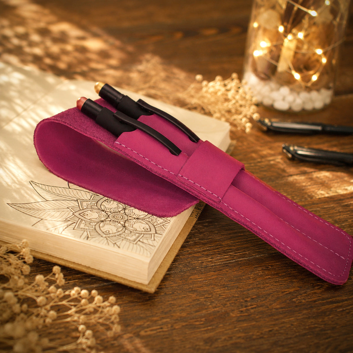 A magenta pink faux-leather pen pouch resting on an open sketchbook on a wooden desk, with two black pens tucked inside. The scene is styled with dried flowers and a warm, glowing glass jar in the background.
