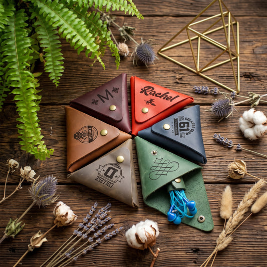 Six small leather card cases arranged in a circle on a rustic wooden surface, each in a different color: dark brown, tan, red, navy, taupe, and sage green. The cases have snap closures, metal studs, and printed monogram or logo designs.