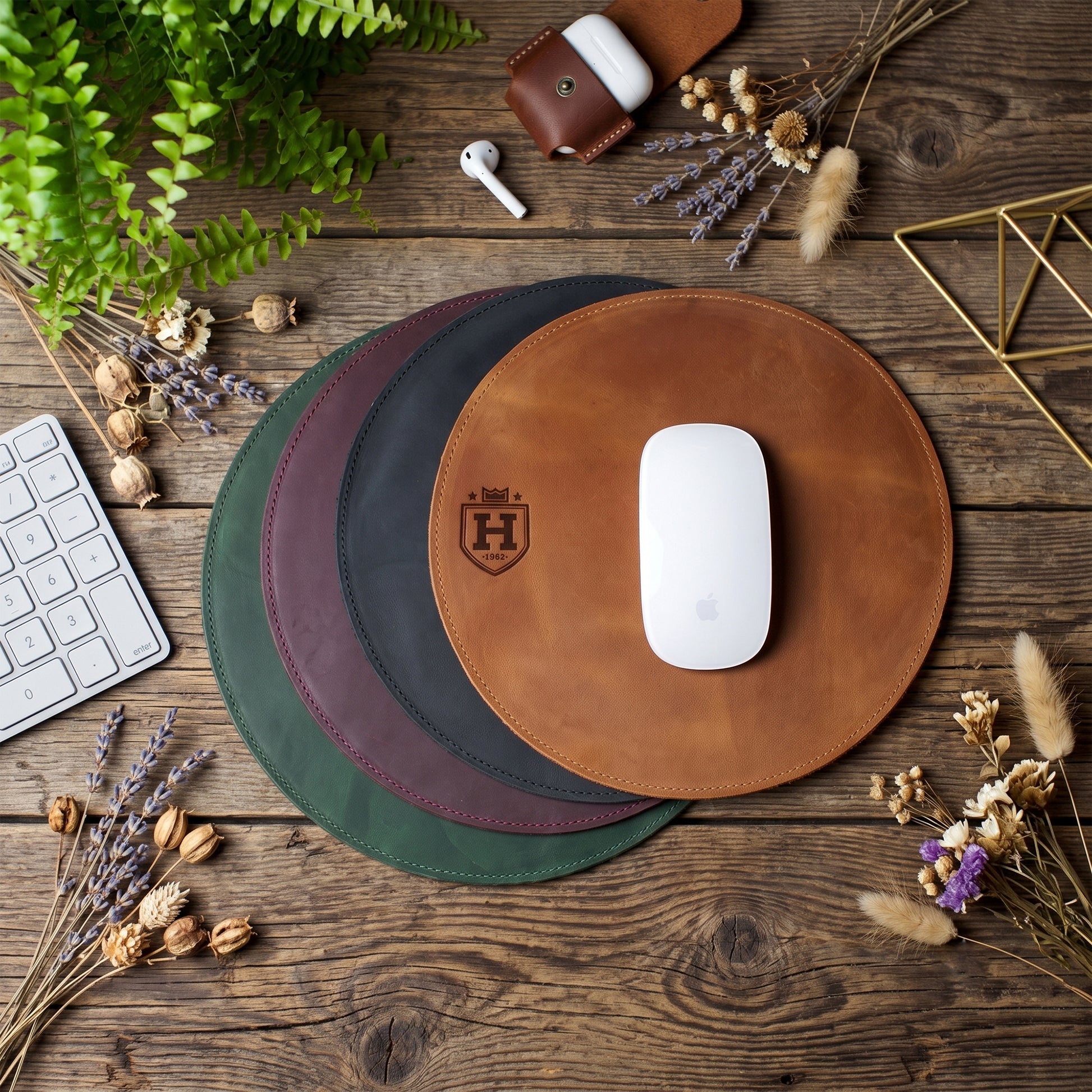 Three round leather mouse pads in brown, dark green, and burgundy are arranged on a rustic wooden desk with a white mouse on the top pad. The scene is styled with dried flowers, a keyboard, a phone case, and other desk accessories.