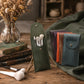 A set of makeup brushes displayed in a green leatherette pouch stand, with several colorful pouches lined up behind it on a rustic wooden table. A glass vase with dried flowers, old books, and a white-handled brush are arranged around the product.