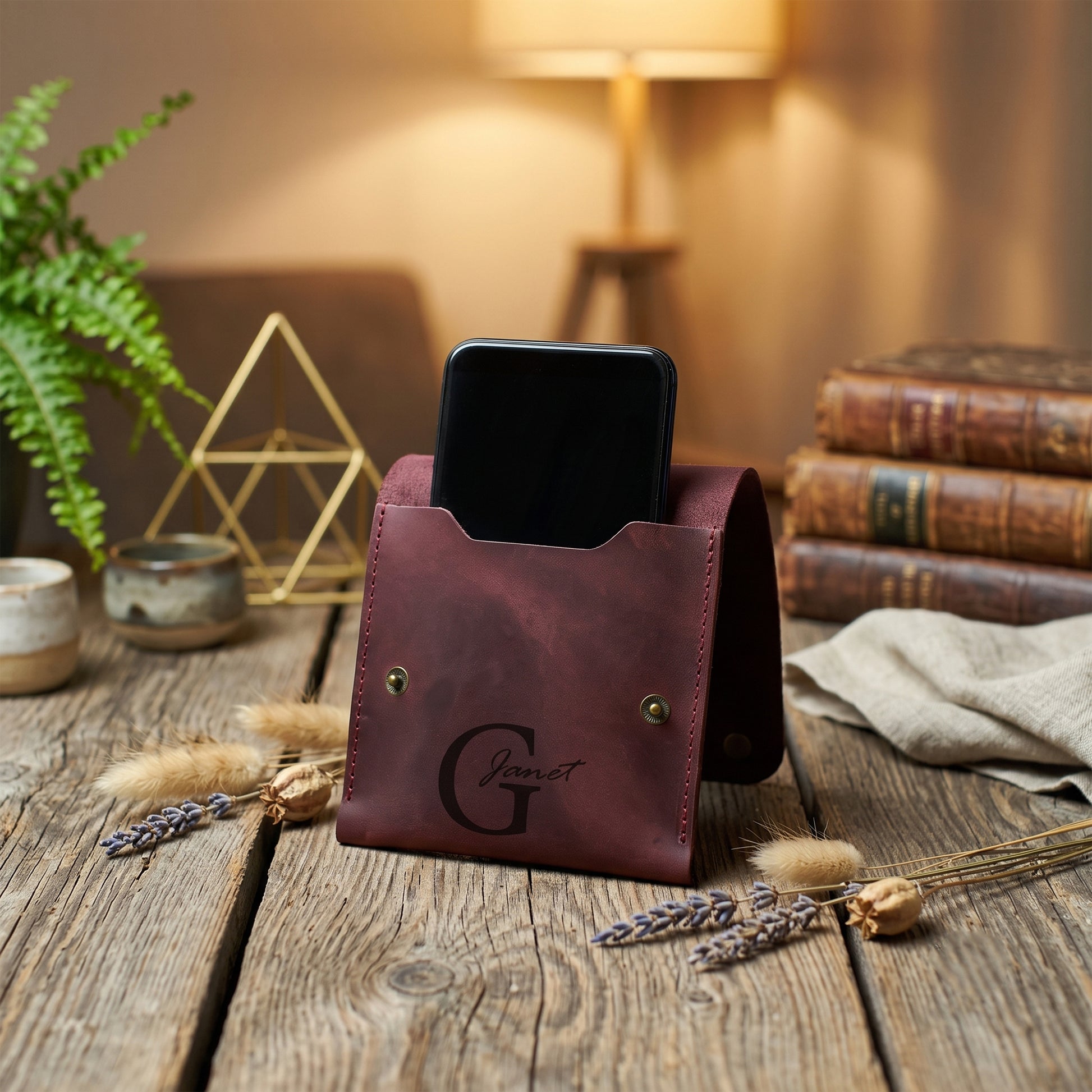 A dark reddish-brown leather phone stand on a rustic wooden table, holding a black smartphone. The stand features a debossed monogram logo and brass-tone rivets, with a warm, cozy background containing books, a lamp, and dried botanicals.