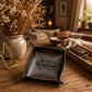 A dark brown leather square tray with 'Welcome' written in black script on the front, styled on a rustic wooden table in a cozy living room setting.